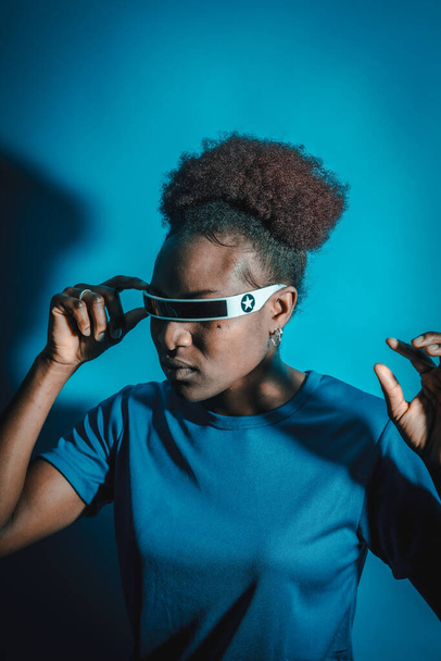 Young African American woman isolated on a blue background with futuristic glasses, studio shoot, future concept - Fotoğraf, Görsel