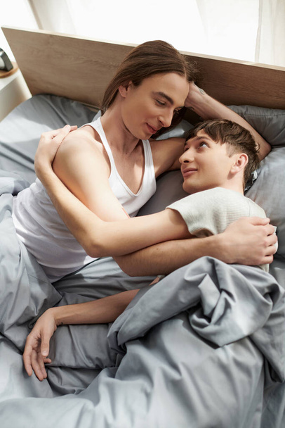 High angle view of young gay couple in pajama hugging and talking with each other  while lying together on comfortable bed in modern bedroom at home in morning  - Foto, Imagen
