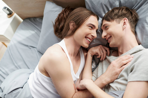 Neşeli ve dövmeli homoseksüel çift sabahları evlerinde, modern yatak odasında birlikte yatarken birbirleriyle konuşuyorlar.  - Fotoğraf, Görsel