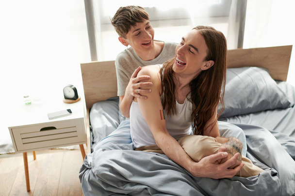 Fröhliches und junges schwules Paar im Pyjama, das Zeit miteinander verbringt und miteinander spricht, während es sich nach dem Aufwachen am Morgen zu Hause auf einem bequemen Bett ausruht  - Foto, Bild