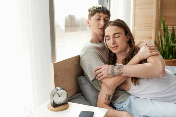 Young gay man with closed eyes hugging tattooed boyfriend in pajama while sitting on bed near alarm clock and mobile phone on bedside table in morning at home  - Φωτογραφία, εικόνα