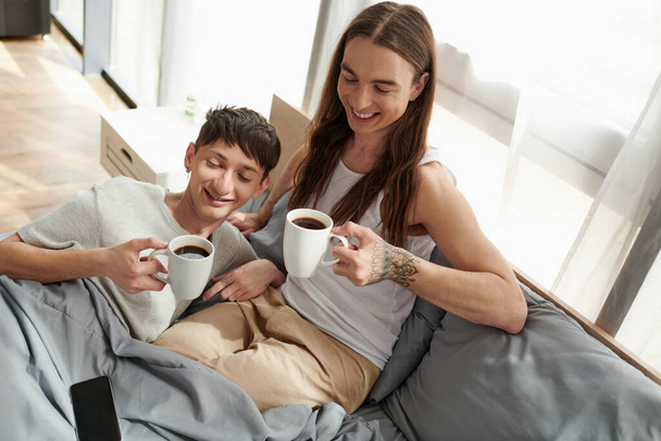 Vista de ángulo alto de la sonriente pareja gay en ropa de dormir sosteniendo tazas de café cerca de teléfono inteligente con pantalla en blanco mientras descansa en la cómoda cama en casa por la mañana  - Foto, Imagen