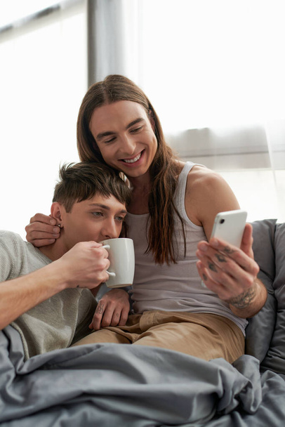 Carefree and long haired homosexual man using smartphone and hugging boyfriend in pajama drinking coffee while sitting together on comfortable bed at home in morning  - 写真・画像