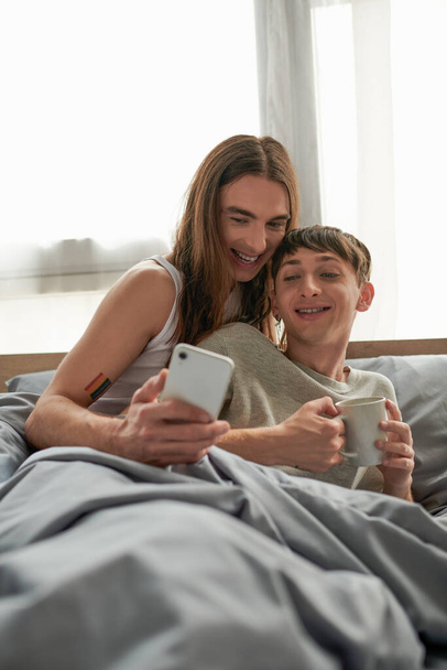 Χαρούμενα μακριά μαλλιά και τατουάζ ομοφυλόφιλος άνδρας χρησιμοποιώντας smartphone δίπλα σε νεαρό φίλο με φλιτζάνι καφέ, ενώ κάθεται κάτω από κουβέρτα στο κρεβάτι στο σπίτι το πρωί  - Φωτογραφία, εικόνα