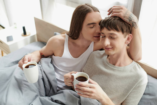 Long haired and tattooed gay man with closed eyes talking and touching head of smiling boyfriend in sleepwear while holding coffee cup on bed in morning at home  - Fotografie, Obrázek