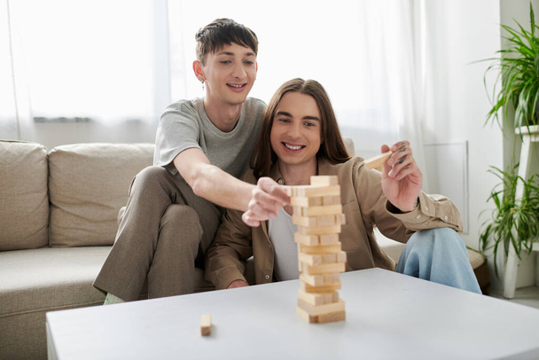 Młoda i szczęśliwa para homoseksualistów w luźnych ubraniach uśmiechnięta podczas gry w drewniane klocki na stole i spędzania czasu razem w nowoczesnym mieszkaniu  - Zdjęcie, obraz