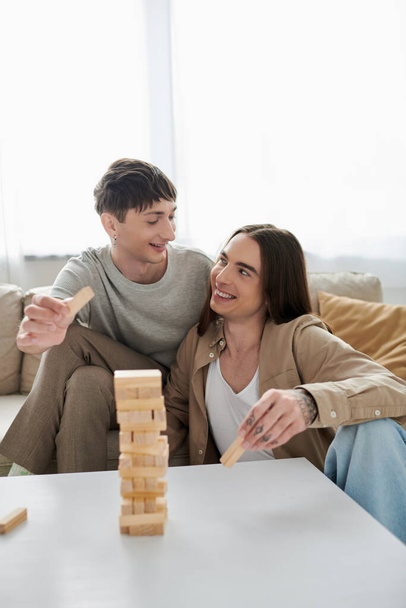 Young and cheerful same sex couple in casual clothes talking and looking at each other while holding parts of wood blocks game near table and couch at home  - Фото, зображення