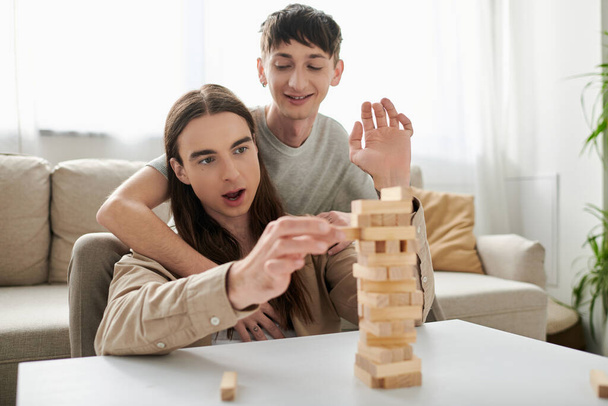 Young and positive gay man hugging long haired boyfriend in casual clothes while playing blurred wood blocks game on table near comfortable couch in living room at home  - Photo, Image