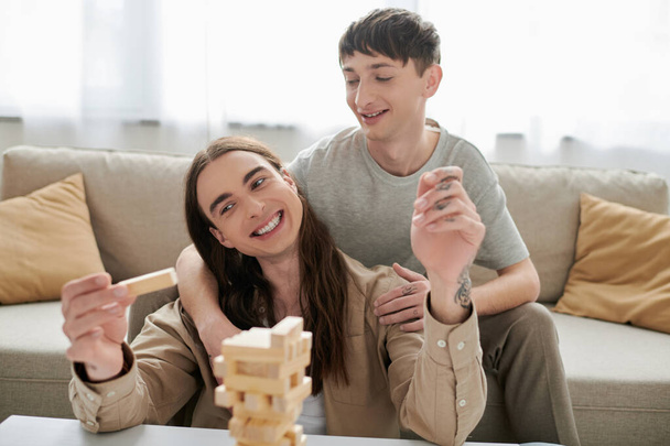 Smiling gay man hugging and looking at long haired and tattooed boyfriend while playing blurred wood blocks game on table near couch in living room at home  - Foto, immagini