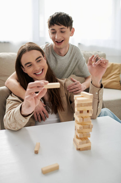 Positive gay man in casual clothes hugging tattooed and long haired boyfriend holding part of blurred wood blocks game on table in living room at home  - Φωτογραφία, εικόνα
