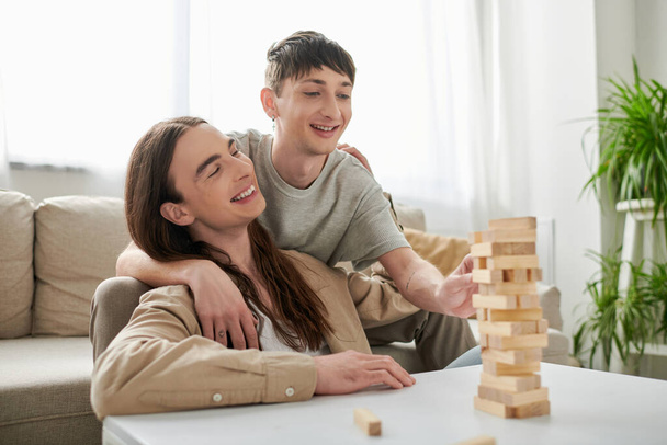 Carefree homosexual man in casual clothes hugging long haired boyfriend and playing wood blocks game on table while spending time together in living room at home  - Foto, Imagem