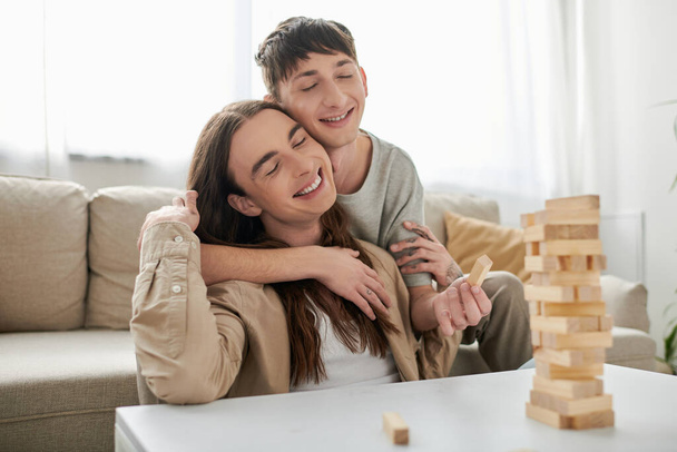 Young smiling gay man with closed eyes hugging long haired boyfriend in casual clothes near blurred wood blocks game on table in living room at home  - Фото, зображення