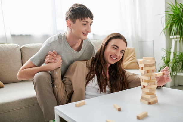 Young and smiling same sex couple in casual clothes holding hands while playing wood blocks game near parts on table in living room at home  - Foto, imagen
