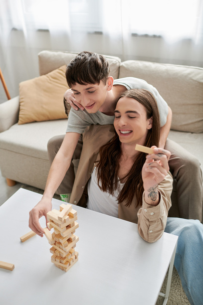 joven gay pareja en casual ropa sonriendo mientras jugando madera bloques juego en mesa cerca cómodo sofá mientras pasando tiempo juntos en sala de estar en casa  - Foto, imagen