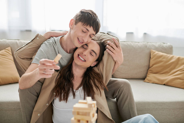 Carefree νεαρό ομοφυλόφιλο ζευγάρι σε casual ρούχα κλείνοντας τα μάτια ενώ αγκαλιάζει και παίζει θολή ξύλο μπλοκ παιχνίδι κοντά στον καναπέ στο σαλόνι στο σπίτι  - Φωτογραφία, εικόνα