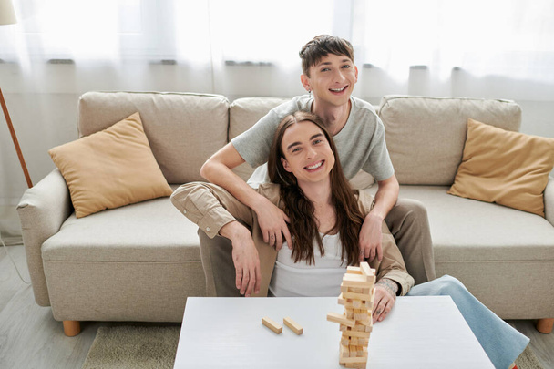 Χαμογελαστό και νεαρό ομοφυλόφιλο ζευγάρι σε casual ρούχα κοιτάζοντας κάμερα κοντά θολή ξύλο μπλοκ παιχνίδι στο τραπέζι, ενώ κάθεται στον καναπέ στο σαλόνι στο σπίτι  - Φωτογραφία, εικόνα