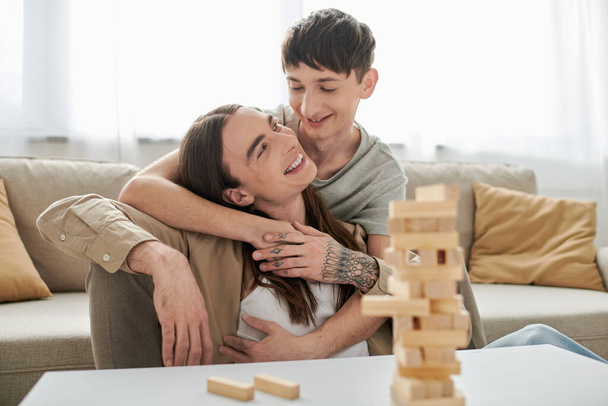 Uśmiechnięty homoseksualista przytulający się i patrzący na długowłosego chłopaka w luźnych ubraniach w pobliżu rozmytych drewnianych klocków gry i części na stole w salonie w domu  - Zdjęcie, obraz