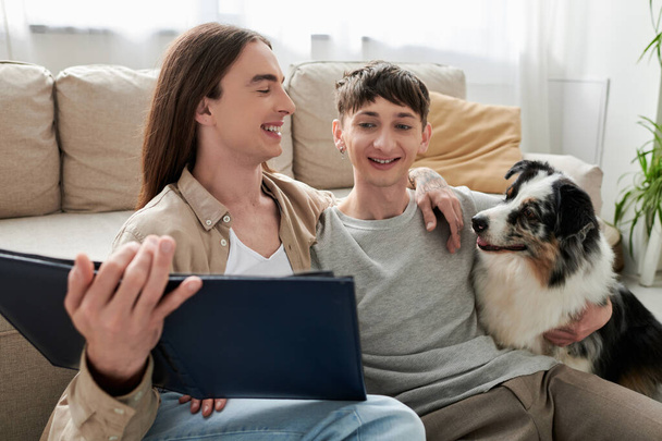 Positive same sex couple in casual clothes looking at blurred photo album and hugging friendly Australian shepherd dog near couch in living room at home  - Photo, Image