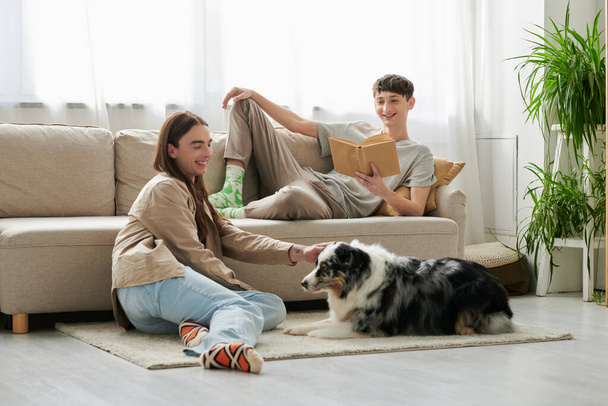 χαρούμενος gay άντρας με μακριά μαλλιά παίζει με αυστραλιανό ποιμενικό σκύλο ενώ ο φίλος του με casual ρούχα ξεκουράζεται στον καναπέ και διαβάζει βιβλίο στο μοντέρνο σαλόνι  - Φωτογραφία, εικόνα