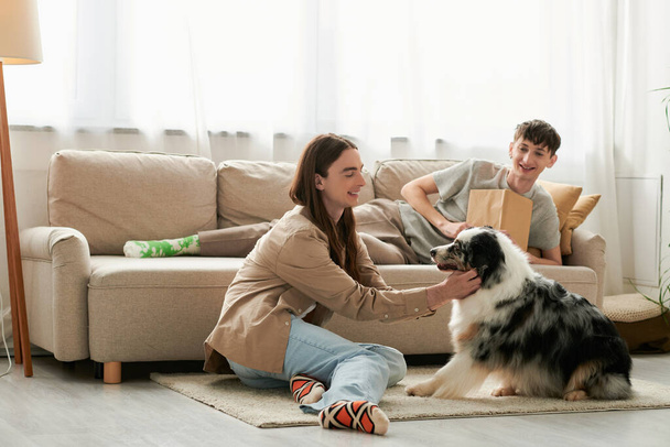 cheerful gay man with long hair playing with Australian shepherd dog while his boyfriend in casual clothes resting on couch and holding book in modern living room  - Foto, imagen
