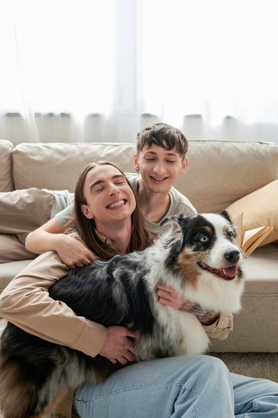 alegre casal lgbt em roupas casuais sentado ao lado de sofá confortável e sorrindo juntos enquanto abraçando cão pastor australiano na sala de estar moderna  - Foto, Imagem