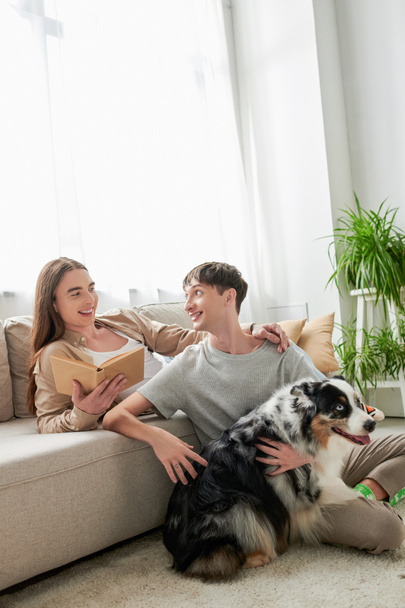 happy gay man with long hair holding book and looking at tattooed and cheerful boyfriend hugging Australian shepherd dog in modern living room at home  - Foto, imagen