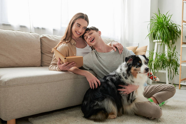 happy gay man with long hair holding book and hugging his tattooed and cheerful boyfriend next to Australian shepherd dog in modern living room at home  - Foto, immagini