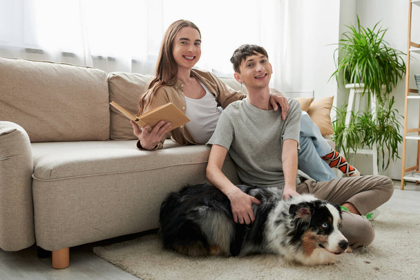 cheerful gay man sitting on carpet and cuddling Australian shepherd dog next to happy partner with long hair holding book and looking at camera in modern living room  - Photo, Image