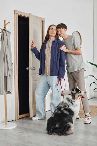 jong gay paar in casual outfits staan met gesloten ogen naast elkaar in hal in de buurt jas rek en Australische herder hond met riem in modern appartement - Foto, afbeelding
