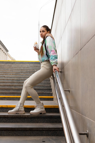 Young Slim brunette girl eating fast food hotdog or burger outdoor on the street on the subway stairs. - Φωτογραφία, εικόνα