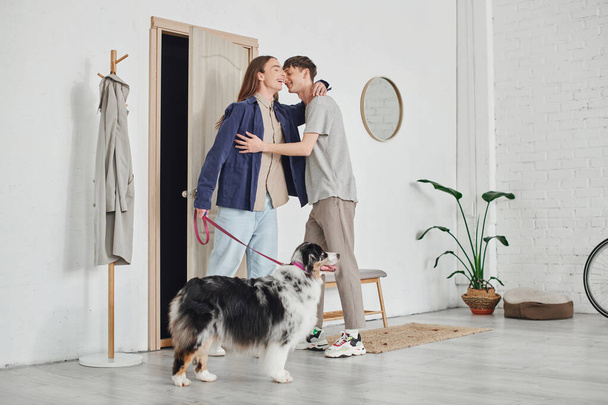 happy gay couple in casual outfits standing and hugging each other in hallway next to coat rack and holding leash near australian shepherd dog while smiling together in modern apartment - Photo, image