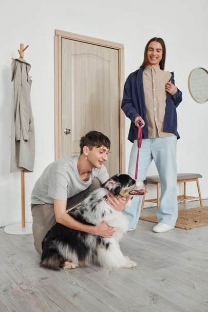 cheerful gay man smiling while cuddling adorable Australian shepherd dog next to joyful boyfriend with long hair holding leash while standing near door in modern hallway  - Foto, afbeelding