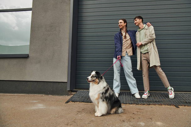 cheerful gay man with pigtails holding leash of Australian shepherd dog and hugging smiling boyfriend in casual outfit while standing together near garage door outside on street - Photo, Image