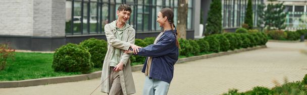 happy lgbt couple holding hands of each other while walking out together and smiling near green bushes and modern building on blurred background on urban street, banner  - Photo, Image