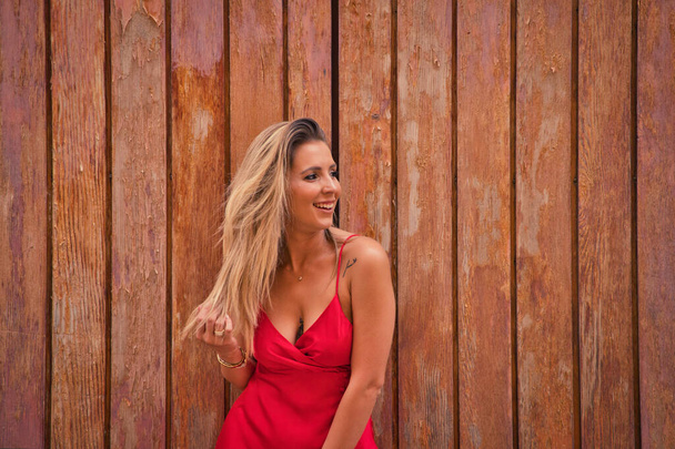 Młoda, piękna kobieta w czerwonej jedwabnej sukience na tle drewnianych drzwi dotyka włosów i sprawia, że różne wyrażenia, patrząc na różne miejsca. Pojęcie wyrażenia i gesty - Zdjęcie, obraz