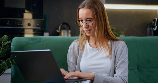 Young woman in glasses sits at on sofa at laptop computer working at home. Female freelancer or student listens to an online conference in the kitchen. - Foto, Imagem