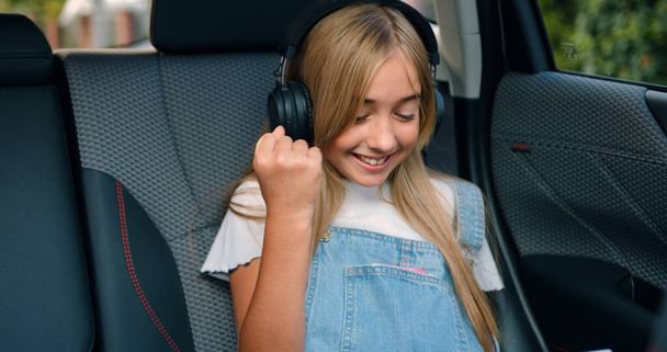 Beautiful positive carefree 12-year-old blond girl in trendy clothes wears wireless earphones searching for something on tablet gadget and reacts emotionally after finding while sitting in autos - Photo, image