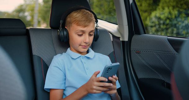 Attractive positive blond 10-year-old boy in wireless earphones applying his mobile for chatting with friends or searching internet for favourite songs,sitting in the car on backseat - Foto, Imagem