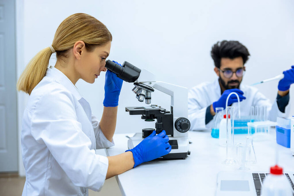 Scientists conducting research investigations in medical laboratory, sitting at table, working together, researcher in foreground using microscope - Foto, Imagem