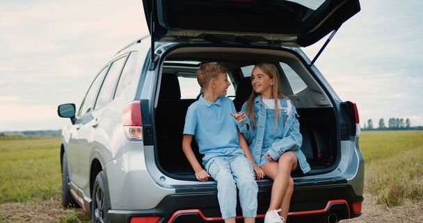 Lovely cheerful amicable 10-12-year-old brother and sister sitting in auto trunk surrounding green lawn and enjoying communication between themselves - Fotoğraf, Görsel