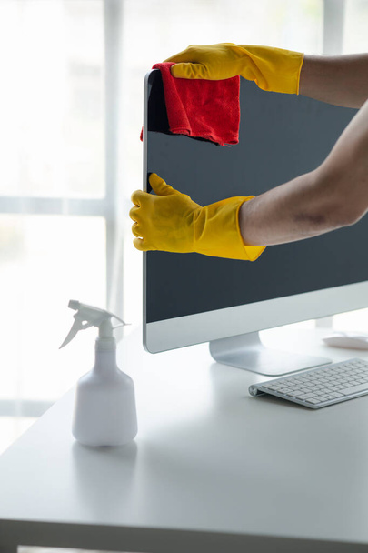 Person cleaning room, cleaning worker is using cloth to wipe computer screen in company office room. Cleaning staff. Concept of cleanliness in the organization. - 写真・画像