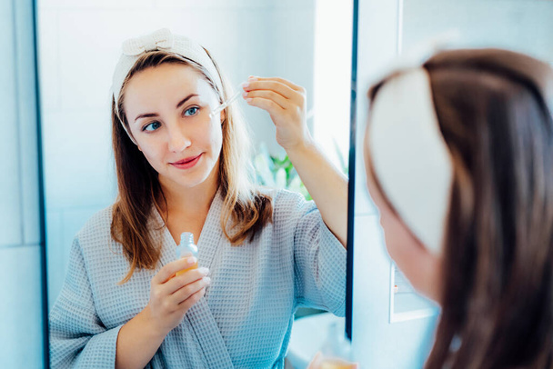 Young woman in bathrobe looking in the mirror, holding dropper with hyaluronic acid serum close to face and smiling. Skin hydrating. Cosmetic spa procedures. Beauty self-care at home. Selective focus - Foto, Imagen
