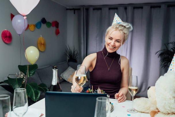 Young woman in party cap drinks champagne and eating cake, having online birthday party with friends. Girl speaking, making video chat, lifestyle blog vlog, webcam view. Stay home, quarantine concept - Фото, изображение