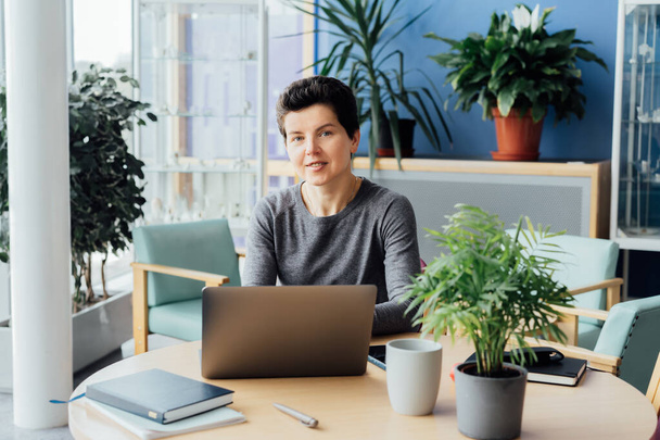 Neutral gender middle aged woman with no make-up in casual clothing using laptop and smiling while working indoors in her workstation in an open space office. Online video call, IT HR, recruiting. - Foto, immagini