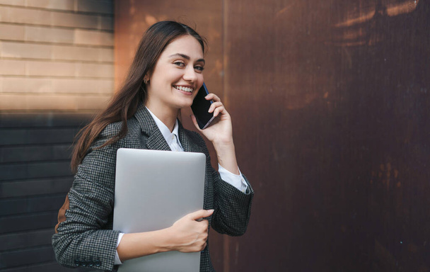 Retrato de uma jovem mulher de negócios sorridente de jaqueta andando pela parte empresarial da cidade com um laptop na mão, falando no telefone celular contra - Foto, Imagem