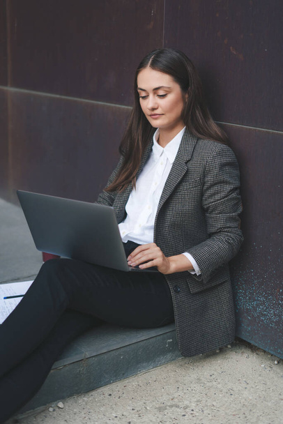 Femme d'affaires réussie vêtue d'une veste assise à l'extérieur et tapant sur un ordinateur portable moderne. Indépendant attrayant utilisant un ordinateur portable pour le travail à distance. Entreprises - Photo, image