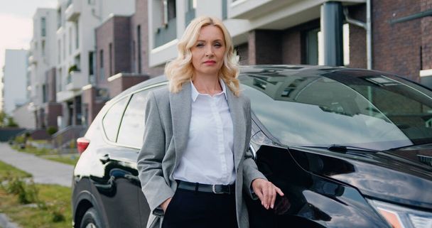 Portrait of successful young caucasian businesswoman in official clothes outside when standing beside black car on parking near modern building. Outdoors. - Fotó, kép