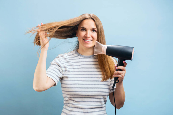Νεαρή χαμογελαστή γυναίκα που χρησιμοποιεί στεγνωτήρα μαλλιών σε μπλε φόντο. Στυλ μαλλιών, έννοια περιποίησης μαλλιών - Φωτογραφία, εικόνα