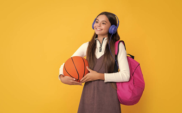 glad child in headphones with school backpack and basketball ball on yellow background. - Foto, afbeelding