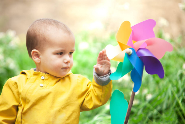 Uma criança pequena com um casaco amarelo brinca com um brinquedo colorido em um prado verde exuberante. O rosto do pequeno está cheio de alegria enquanto o vento gira o volante e eles desfrutam do belo dia ao ar livre. - Foto, Imagem